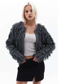 Grå fransjacka med en texturerad, ruggig design. Har dragkedja framtill och framträdande fransdetaljer på ärmarna. Lager över en vit topp.