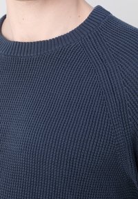 Granatowy sweter z dzianiny o teksturowanym ribbingu. Posiada okrągły dekolt oraz rękawy raglanowe. Materiał jest miękki i elastyczny.