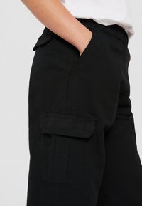 Pantalons cargo noirs en tissu texturé. Comprend une taille élastique, des poches latérales et une poche à rabat sur la jambe.