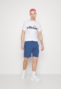 Hvit t-skjorte med svart logo, blå shorts med snor, og hvite joggesko med synlige snørebånd og strukturert stoff; modellen står mot en ensfarget bakgrunn.
