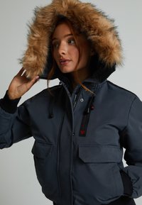 Pelēks ziemas mētelis ar ādas apdari ap kapuci, tam ir rāvējslēdzis priekšā, divas priekšējās kabatas, melni akcenti un regulējami auklas.