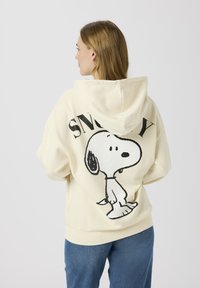 Felpa con cappuccio color crema con tasca a marsupio frontale, caratterizzata da una grande grafica nera di Snoopy e dal testo "SNOOPY" sul retro. Tessuto morbido.