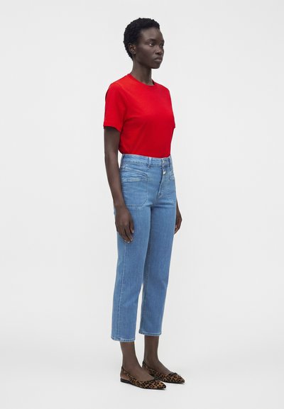 Femme portant un t-shirt rouge, un jean court taille haute bleu clair et des ballerines pointues imprimé léopard devant un fond uni.
