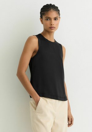 Młoda kobieta z zaplecionymi włosami, ubrana w czarny, bezrękawnik i kremowe spodnie z prążkowanego sztruksu, stoi na tle białej ściany.