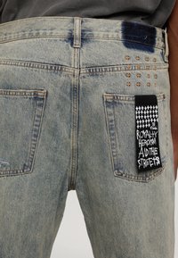 Jean en denim bleu clair avec deux poches arrière, des clous dorés en forme de croix près de la poche droite, et une étiquette en tissu noire avec un texte blanc attachée.