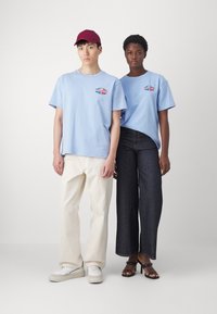 Camisetas de algodón azul claro con logo, combinadas con pantalones color crema y jeans oscuros. Uno lleva una gorra bordó y el otro sandalias negras.