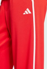 Crvene atletske hlače od glatkog materijala imaju bijele vertikalne pruge i bijeli Adidas logo na gornjem lijevom bedru.