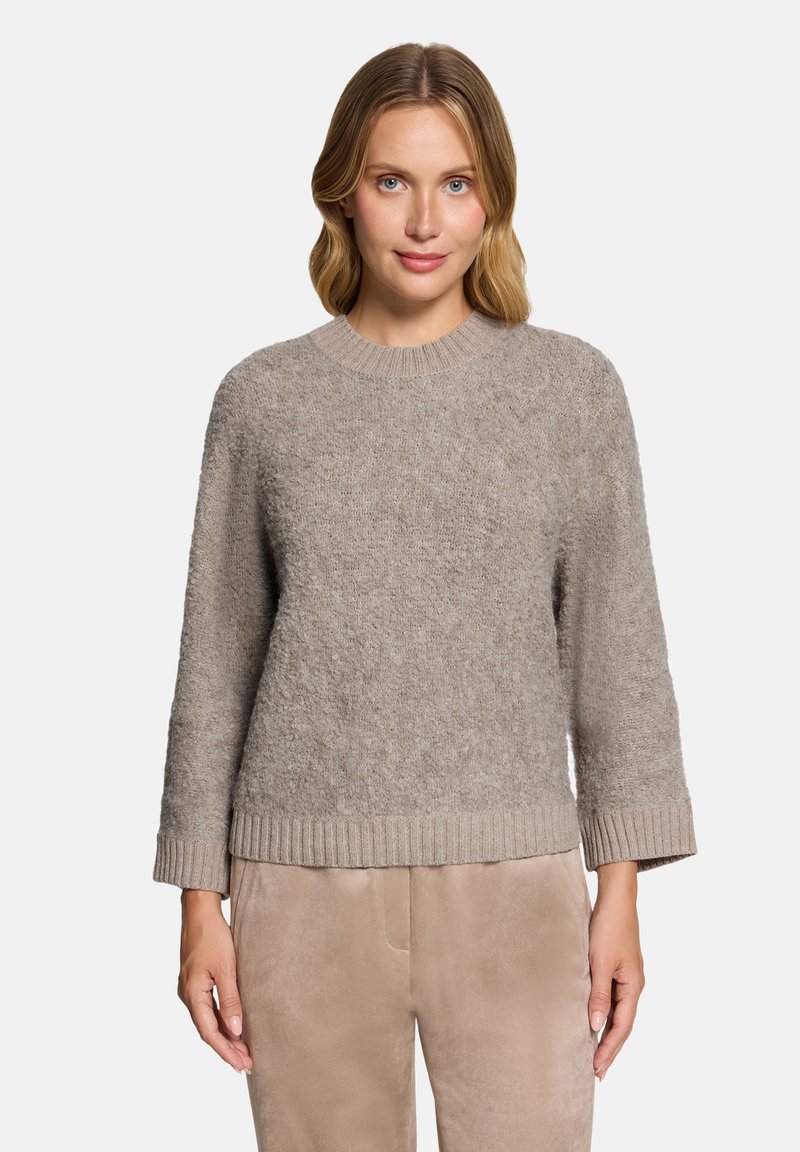Femme aux cheveux blonds mi-longs portant un pull texturé marron clair et un pantalon beige, debout devant un fond clair uni.
