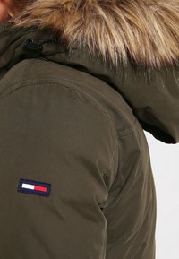 Olive green winter jacket with a fur-trimmed hood and a rectangular logo patch featuring red, white, and navy blue stripes.
