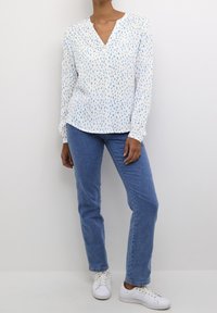 Chemise blanche à manches longues avec un motif bleu et jaune, col en V, associée à un jean bleu et des baskets blanches. Texture de tissu lisse.