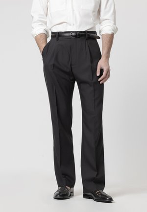 Hombre de pie con las manos en los bolsillos, vistiendo pantalones negros plisados, cinturón negro, camisa blanca con las mangas remangadas y zapatos de vestir negros