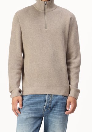 Mężczyzna ubrany w beżowy sweter z zamkiem do połowy długości i niebieskie, wyblakłe dżinsy, stojący z rękami swobodnie opuszczonymi wzdłuż ciała na białym tle.