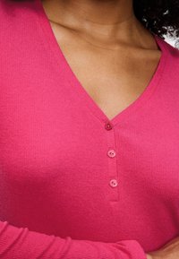 Roze geribde cardigan met een V-halsontwerp, voorzien van vier bijpassende knopen aan de voorkant en lange mouwen. Zachte textuur.