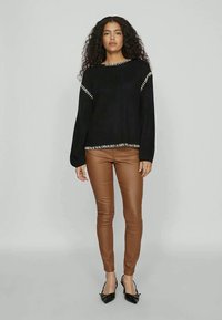 Čierny rebrovaný sveter s krémovými akcentmi, skombinovaný s priliehavými hnedými koženými nohavicami. Modelka má na sebe čierne podpätky a stojí na neutrálnom pozadí.