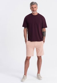 T-shirt en coton bourgogne avec un col rond, associé à un short rose clair. Le modèle porte des baskets beiges et se tient devant un fond neutre.