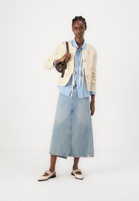 Beež tepitud jakk lilleliste tikanditega, helesinine kraega särk, pikk denim seelik ja beežad lapikud kingad rihmade detailidega, hoides tumedat kotti.