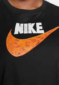 Černé tričko s bílým logem "NIKE" a oranžovým swoosh, vyrobené z měkké bavlny s kulatým výstřihem.