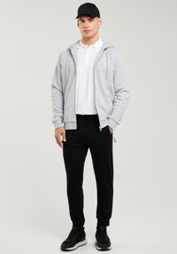 Mees, kes kannab musta nokamütsi, helehallit lukuga kapuutsi, valget polo, musti dressipükse ja musti spordijalatsid, seisab ühetaolise hele tausta ees.