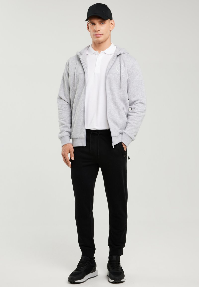 Mees, kes kannab musta nokamütsi, helehallit lukuga kapuutsi, valget polo, musti dressipükse ja musti spordijalatsid, seisab ühetaolise hele tausta ees.