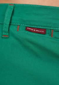 Tessuto di cotone verde con texture visibile, caratterizzato da cuciture rosse e un'etichetta bordeaux che recita "CIPO & BAXX." Primo piano dell'area della vita.