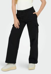 Mustat cargo-housut, joissa on leveä lahjesuunnittelu, joustava vyötärö ja sivutaskut, valmistettu pehmeästä kankaasta, yhdistetty valkoisiin lenkkareihin.