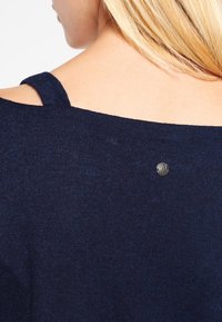 Haut en maille bleu marine avec une sangle au dos et un petit accent métallique rond. Texture lisse avec une coupe légèrement ample, idéale pour être superposée.