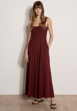 Kobieta stojąca w bordowej, bezrękawnikowej sukni sięgającej podłogi z marszczonym gorsetem i szerokimi ramiączkami, w zestawie z ciemnobrązowymi sandałami.