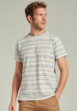 Man with short curly hair wearing a beige striped short-sleeve shirt and tan pants, looking to the right with hand in pocket.