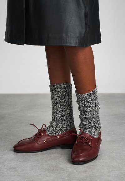 Bordeauxrote Lederschuhe mit goldenen Ösen, kombiniert mit dicken, strukturierten grauen und schwarzen Rippstrumpfsocken. Vor einem neutralen Hintergrund.