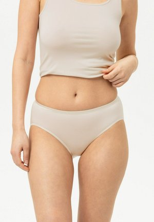 Femme portant des sous-vêtements beige sans coutures et un débardeur assorti, tenant l’ourlet du haut d’une main sur fond blanc.
