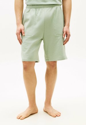 Persoon die lichtgroene knielange shorts met voorzakken draagt, blootsvoets staand op een effen witte achtergrond.