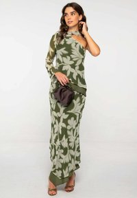 Vestido largo verde oliva con estampados florales, diseño de un solo hombro y tela fluida. Combinado con un bolso pequeño marrón y zapatos de punta abierta.