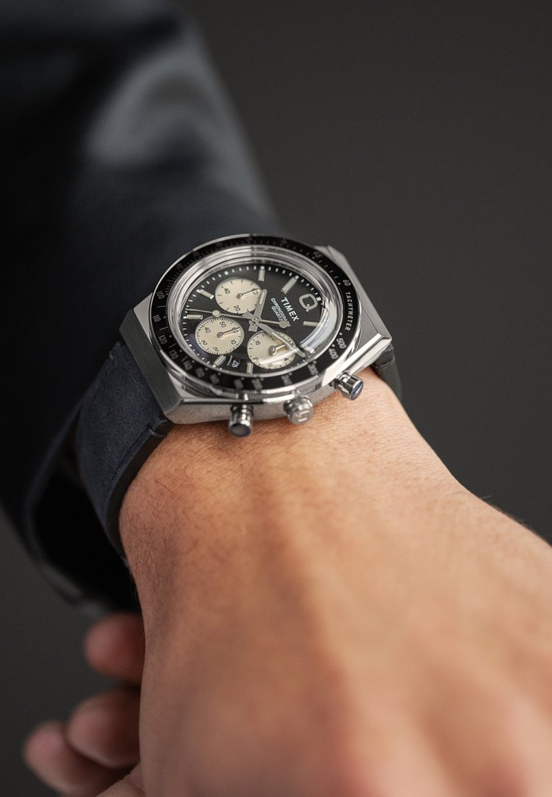 Montre chronographe avec un cadran noir, un boîtier en métal argenté et trois sous-cadrans. Présente un bracelet noir et des accents en acier inoxydable.