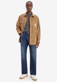 Giacca camicia in corduroy marrone con tasche sul petto, indossata sopra una t-shirt grigia, abbinata a pantaloni jeans blu a gamba dritta e sneakers bianche.
