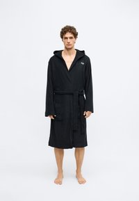 Robe de banho preta com capuz, feita de tecido de felpa texturizado, apresentando um cinto na cintura e dois bolsos frontais, com um logótipo da marca visível no peito.