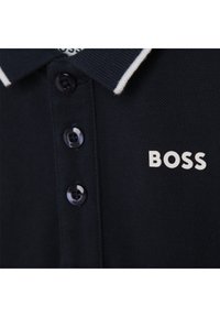 Sötétkék póló texturált anyagból, fehér szegélyes gallérral, három sötét gombbal és a mellkason jól látható, fehér "BOSS" logóval.