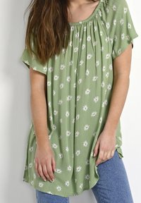 Zielona kwiecista bluzka wykonana z lekkiego materiału, z marszczonym dekoltem i wzorem białych stokrotek na całej powierzchni. W zestawie z niebieskimi dżinsami.