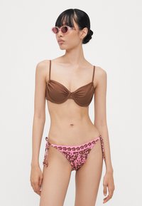 Rudas paminkštintas bikini viršus su plonais dirželiais; rožinė gėlių rašto bikini apačia su reguliuojamomis raišteliais. Modelis nešioja rožinius akinius nuo saulės.