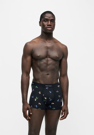 Homme musclé à la peau foncée debout, portant un boxer noir avec des motifs colorés de cerises sur un fond blanc.