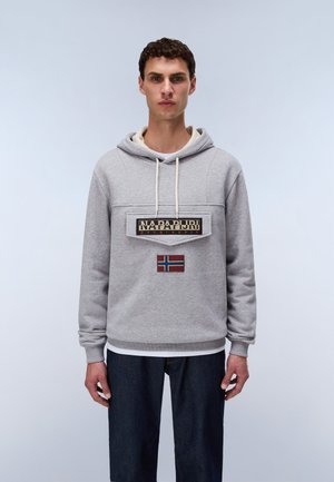 Jeune homme portant un sweat à capuche gris Napapijri avec une poche frontale et un écusson du drapeau norvégien, associé à un jean bleu foncé.