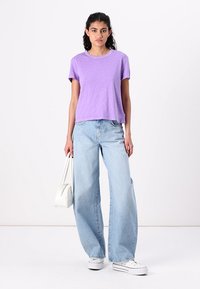 Lavendel kortermet T-skjorte med en avslappet passform, sammen med lyseblå vide jeans og hvite kanvas sneakers. Hvit håndveske til stede.