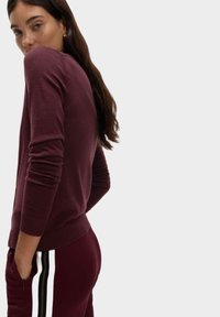 Kobieta z długimi brązowymi włosami, ubrana w bordowy sweter i pasujące spodnie z białymi i czarnymi bocznymi paskami, stojąca na jednolitym tle.