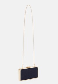 Suuri tummansininen clutch-laukku, jossa on kultakehys ja pitkä ohut kultaketju, roikkuu pystysuorassa valkoista taustaa vasten.
