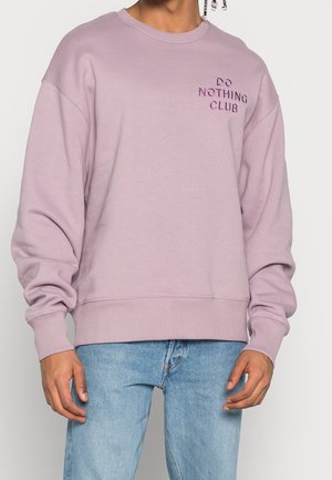 Person trägt einen hellvioletten Pullover mit dem Aufdruck "DO NOTHING CLUB" auf der Brust, kombiniert mit hellblauen Jeans.