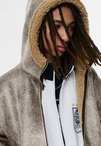 Veste zippée marron avec une doublure de capuche en fourrure beige, présentant une couche intérieure blanche avec un imprimé graphique et des étoiles. Tissu texturé avec un aspect usé.