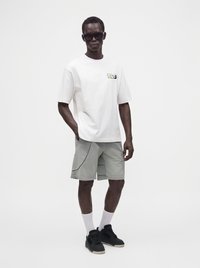 Man wearing white graphic t-shirt, gray shorts with stitched details, white crew socks, black sneakers, and dark sunglasses standing against plain background.