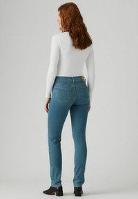 Kape skinny biru dari denim dengan pinggang tinggi, dilengkapi dengan sepasang kantong belakang dan detail jahitan yang halus, dipadukan dengan atasan putih berlengan panjang.