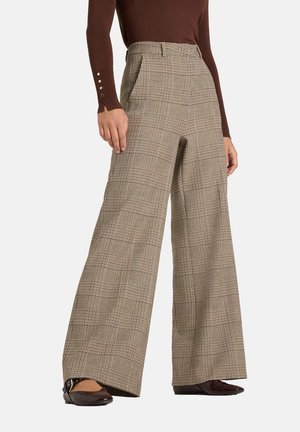 Frau trägt karierte weit geschnittene Hosen, ein dunkelbraunes Langarmshirt mit Knopfdetails und dunkelbraune spitze Schuhe.