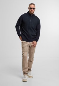 Marineblauer Reißverschlusspullover mit hohem Kragen, beiges Hosen und weiße Sneaker. Das Model steht selbstbewusst vor einem hellen Hintergrund.