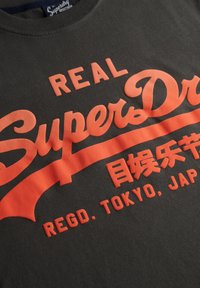 Camiseta negra de Superdry con texto en relieve de color naranja que dice "REAL SuperDry", caracteres japoneses y "REGD. TOKYO, JAPAN."